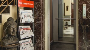 A marble-floored lobby with a Buddha statue, a staircase with a decorative railing, and a newspaper rack with 'The Times of India' and 'Education Times' magazines. - Hotel Sita International (New Delhi)