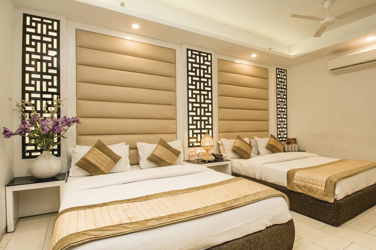 Habitación de hotel con dos camas, cabeceros color beige, paneles decorativos, una mesita de noche con lámpara y un jarrón con flores.