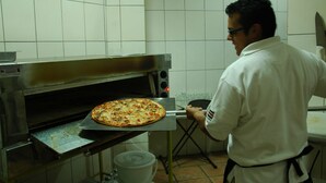 Cuina amb forn de pizza, una pizza que s'està traient i una persona uniformada de cuiner.