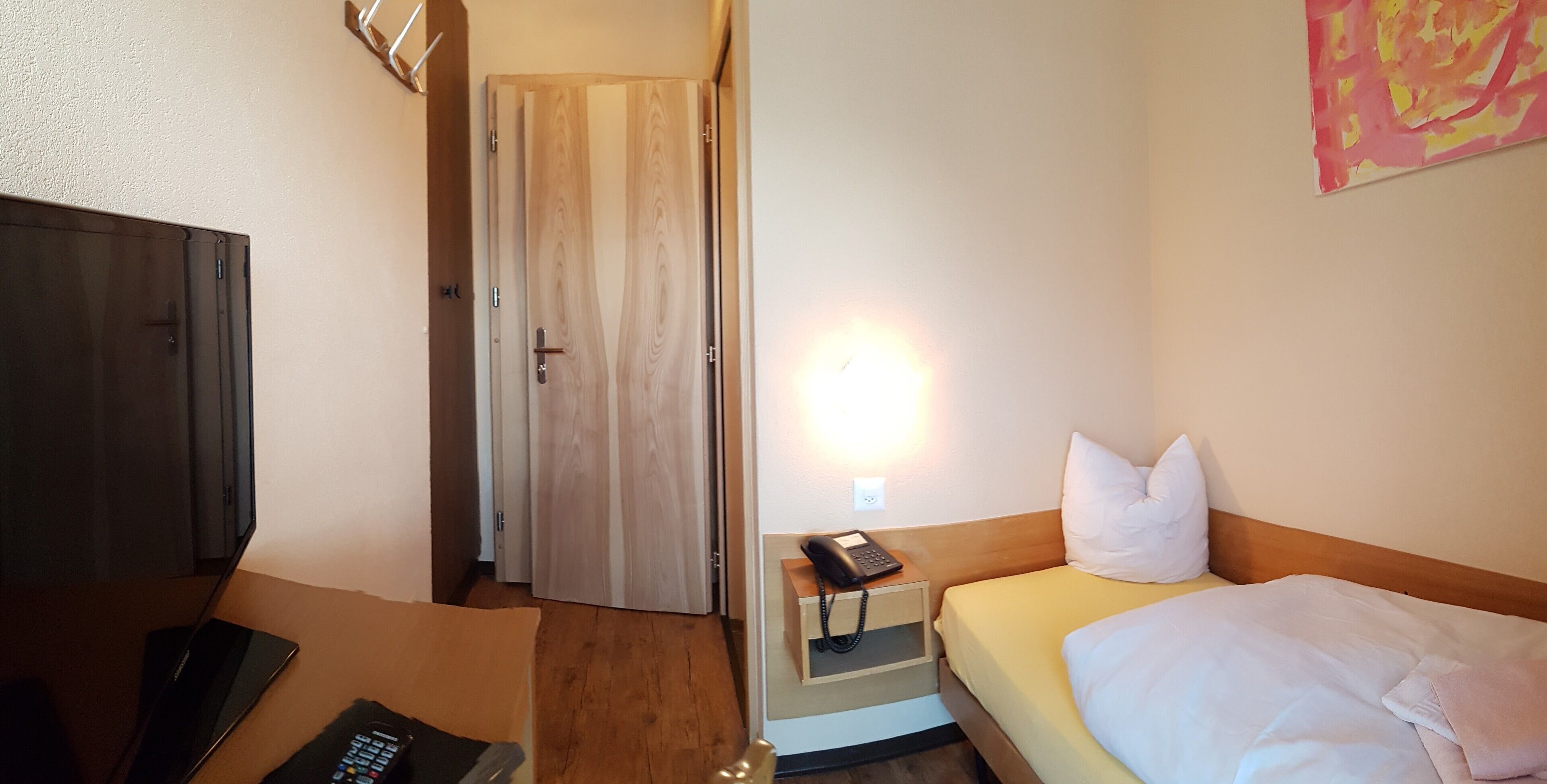 A hotel room with a bed, a bedside table with a phone, a TV, and a door leading to another room.