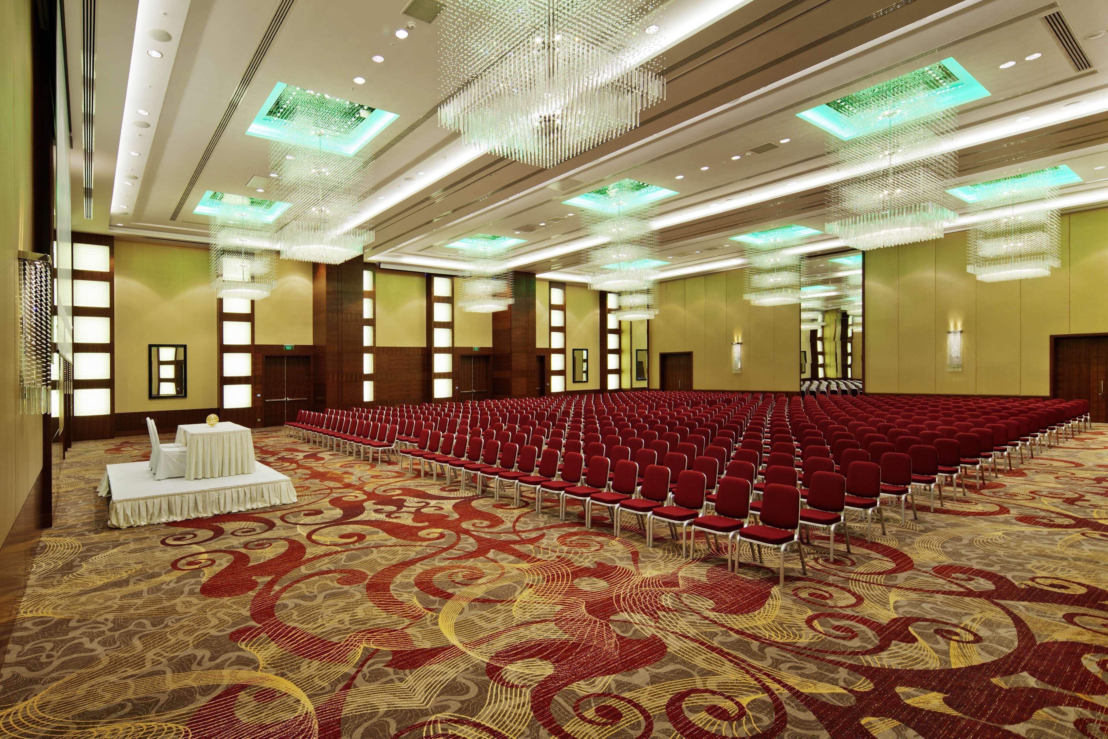A large conference room with rows of red chairs, a patterned carpet, and modern chandeliers.