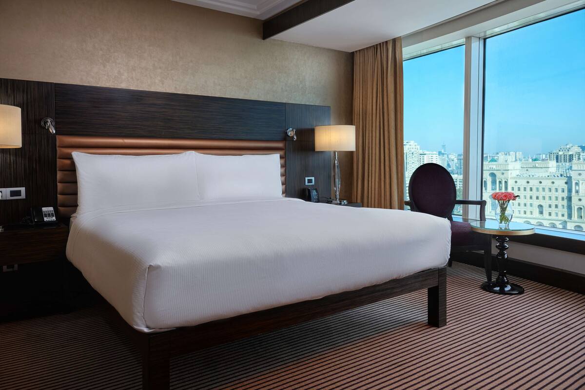 Habitación de hotel con una cama grande, dos lámparas de noche, una mesita de noche con un teléfono y vistas a la ciudad a través de amplios ventanales.