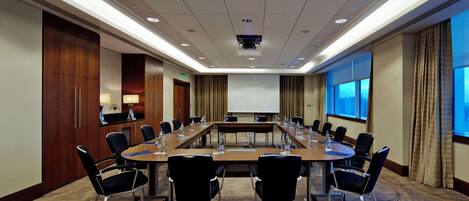 Un salón de conferencias con una larga mesa, sillas, una pantalla de proyector y una pantalla de presentación.