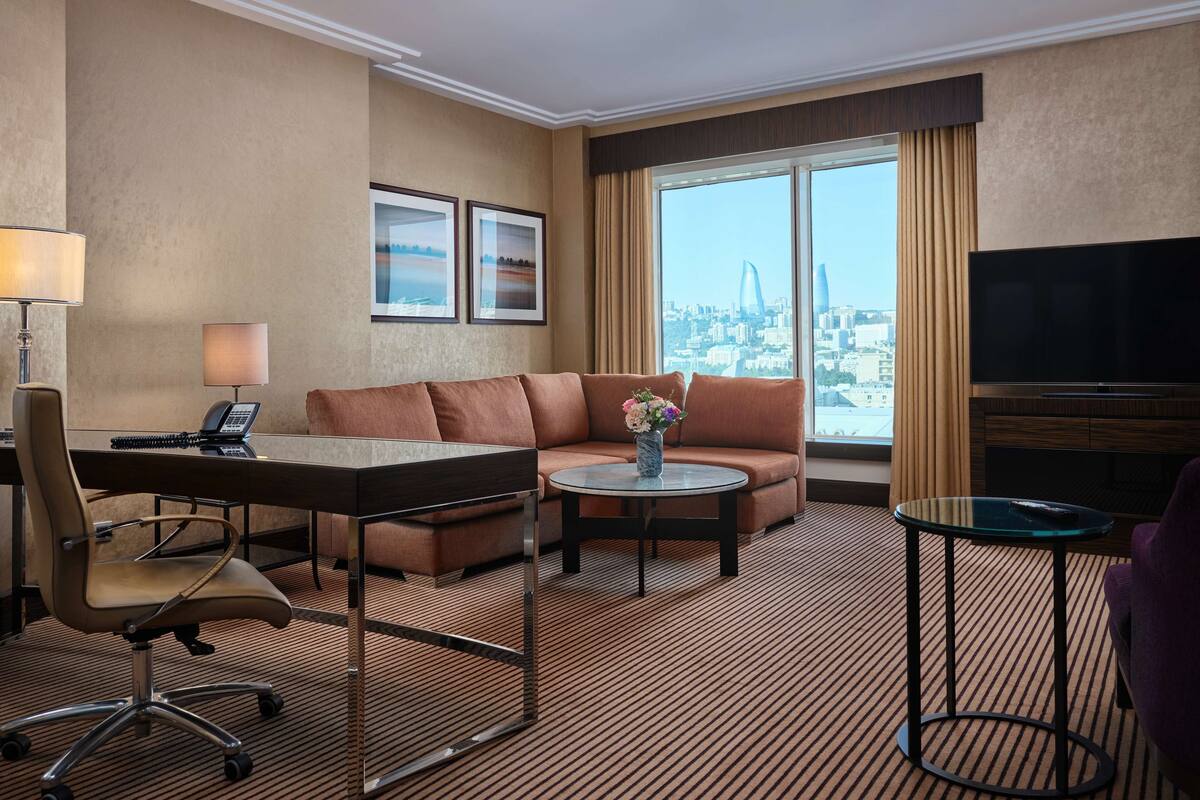 Habitación de hotel con sofá, escritorio con teléfono, una mesita con un jarrón, televisión y vistas a la ciudad a través de amplias ventanas.