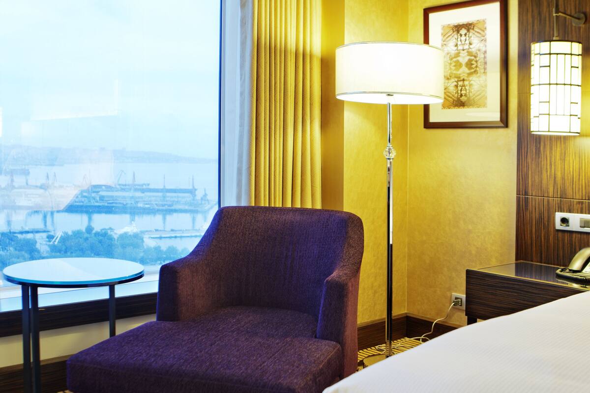 Habitación de hotel con una cama grande, un sillón púrpura, una mesita pequeña, una lámpara de pie y vista a un paisaje urbano a través de un ventanal grande.