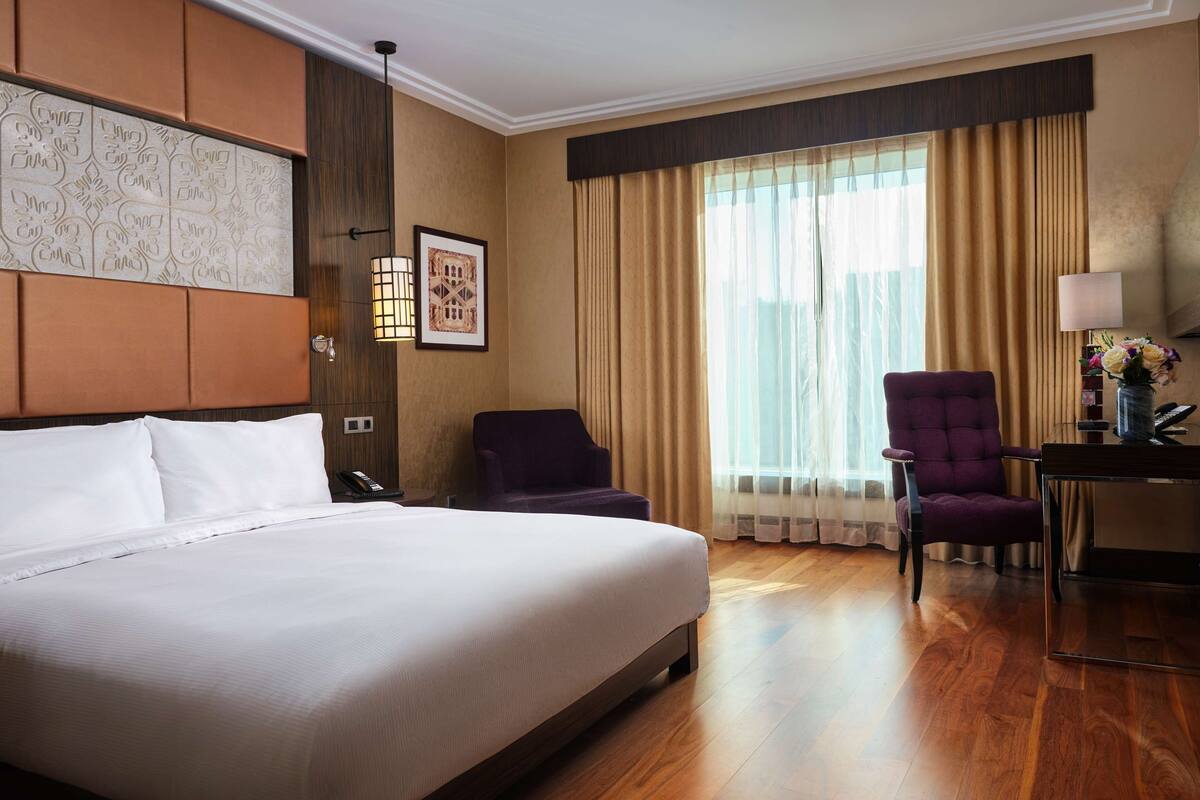 Una habitación de hotel con una cama grande, dos sillones, un escritorio con lámpara y una ventana con cortinas.