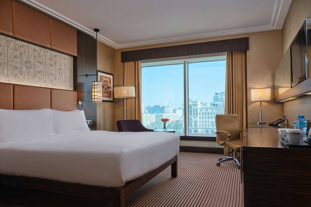 Habitación de hotel con una cama grande, un escritorio con lámpara, una silla y vistas a la ciudad a través de amplias ventanas.