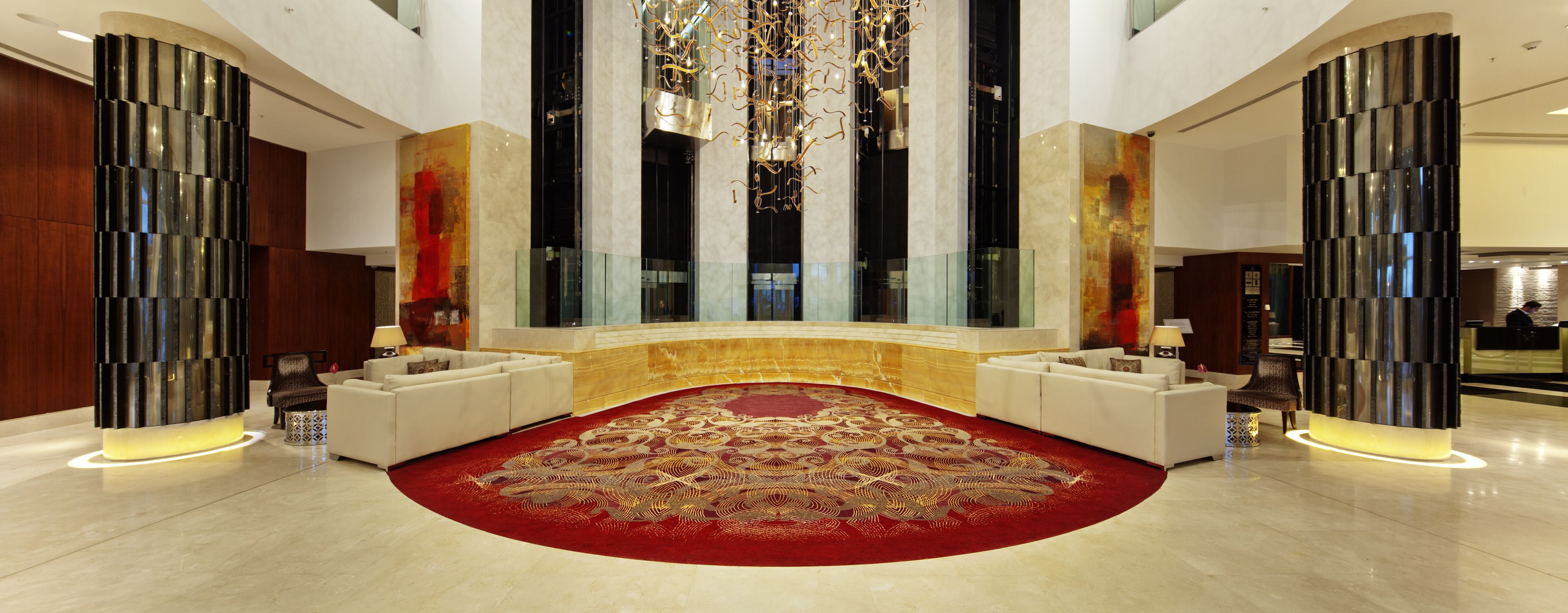 A spacious lobby with a red patterned carpet, two white sofas, a large chandelier, and modern decor.
