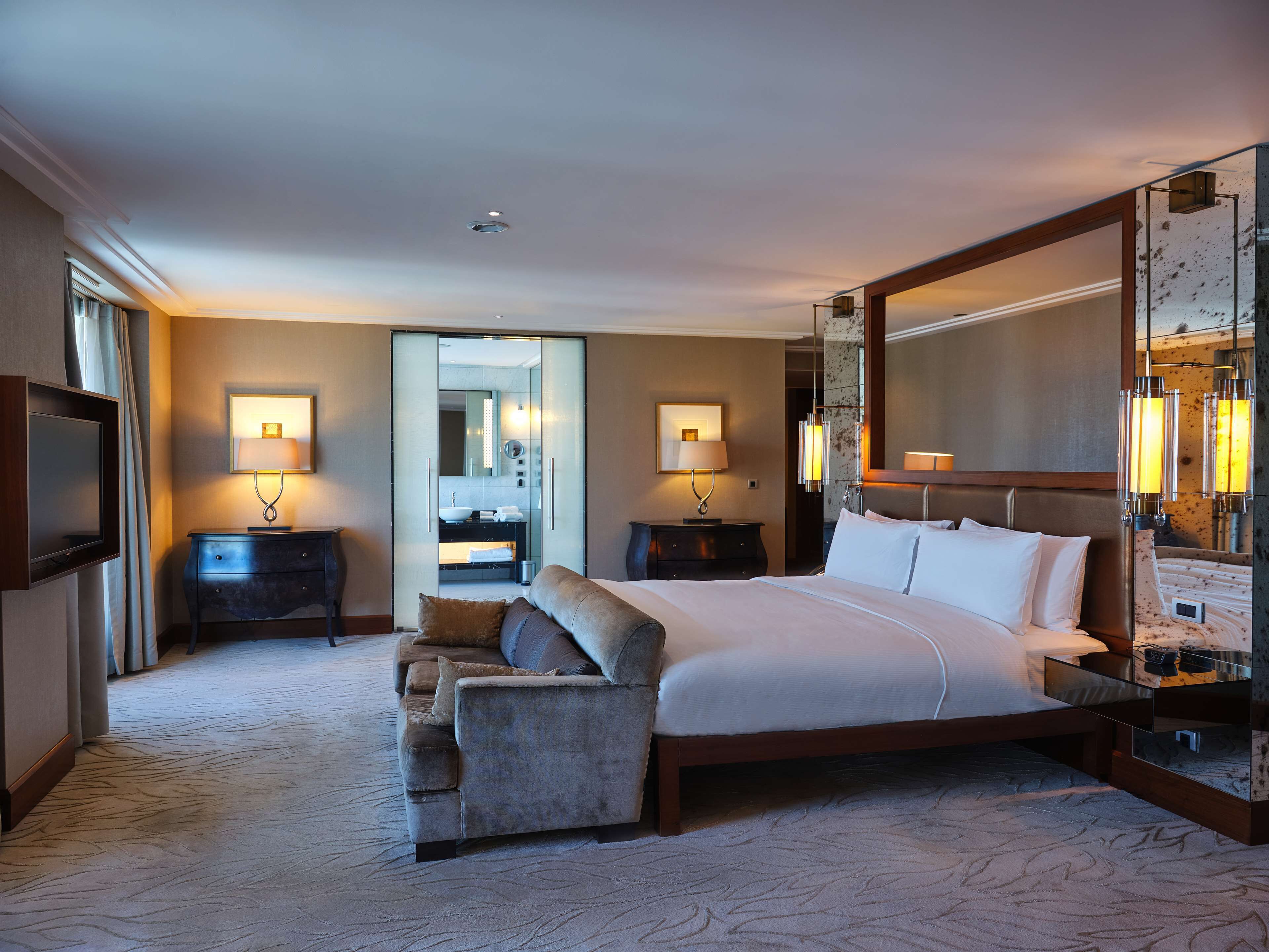 Ein Hotelzimmer mit einem großen Bett, einer Sitzecke, einem Fernseher, einem Schreibtisch und einem durch eine Glastür einsehbaren Badezimmer.