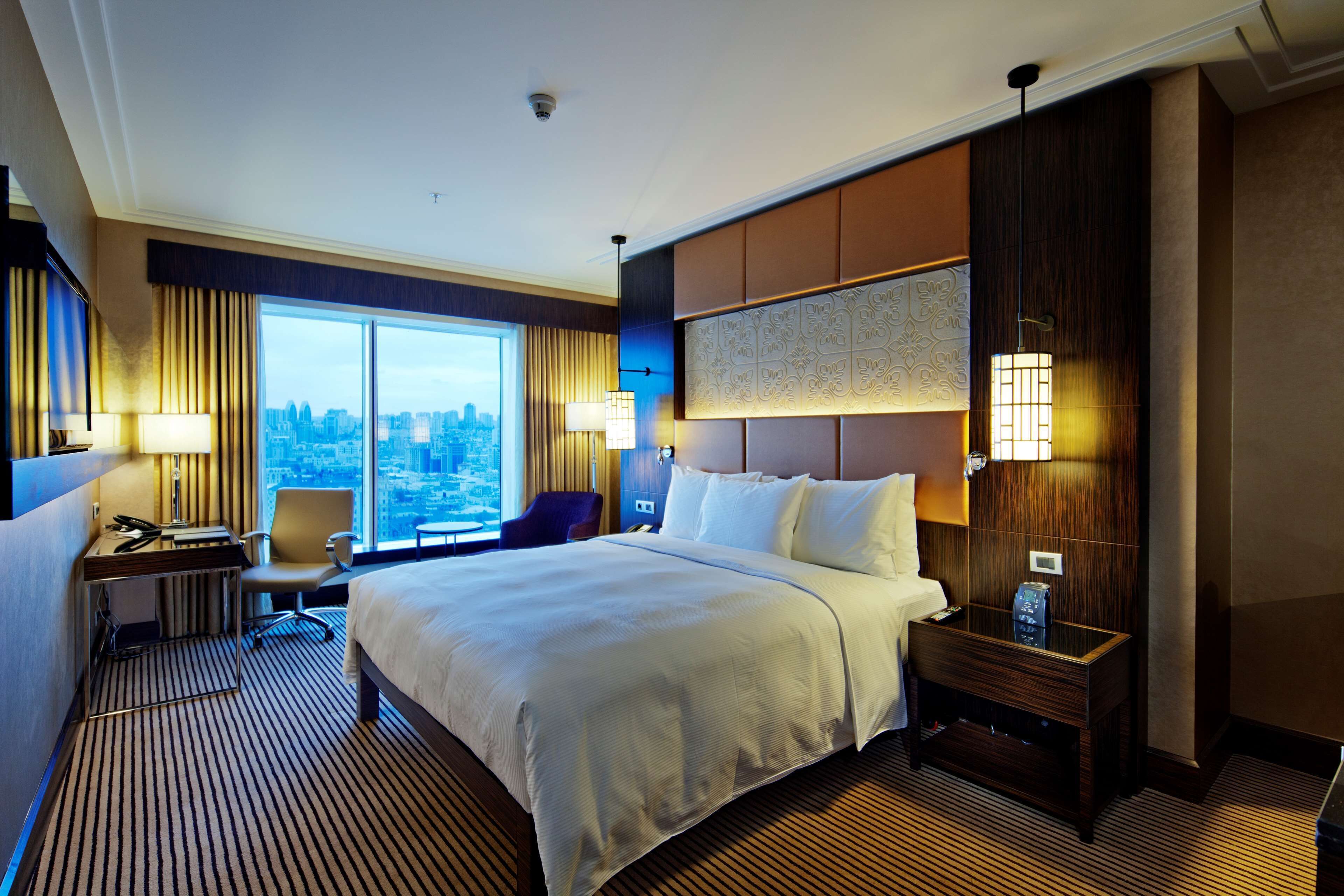 A hotel room with a large bed, a desk with a chair, a nightstand, and a view of the city through large windows.