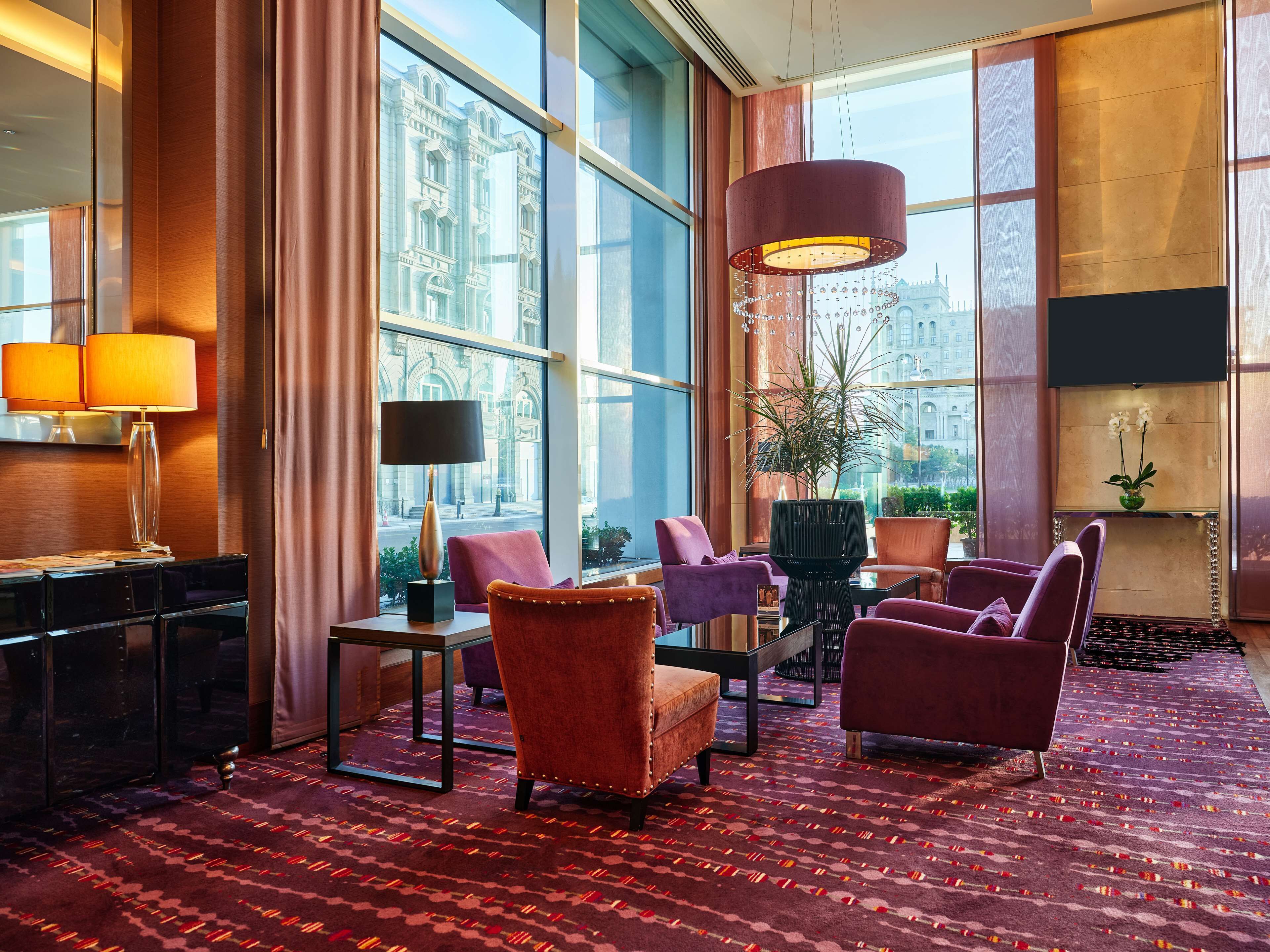 A hotel lobby with large windows, a red patterned carpet, and modern furniture including armchairs and a table.