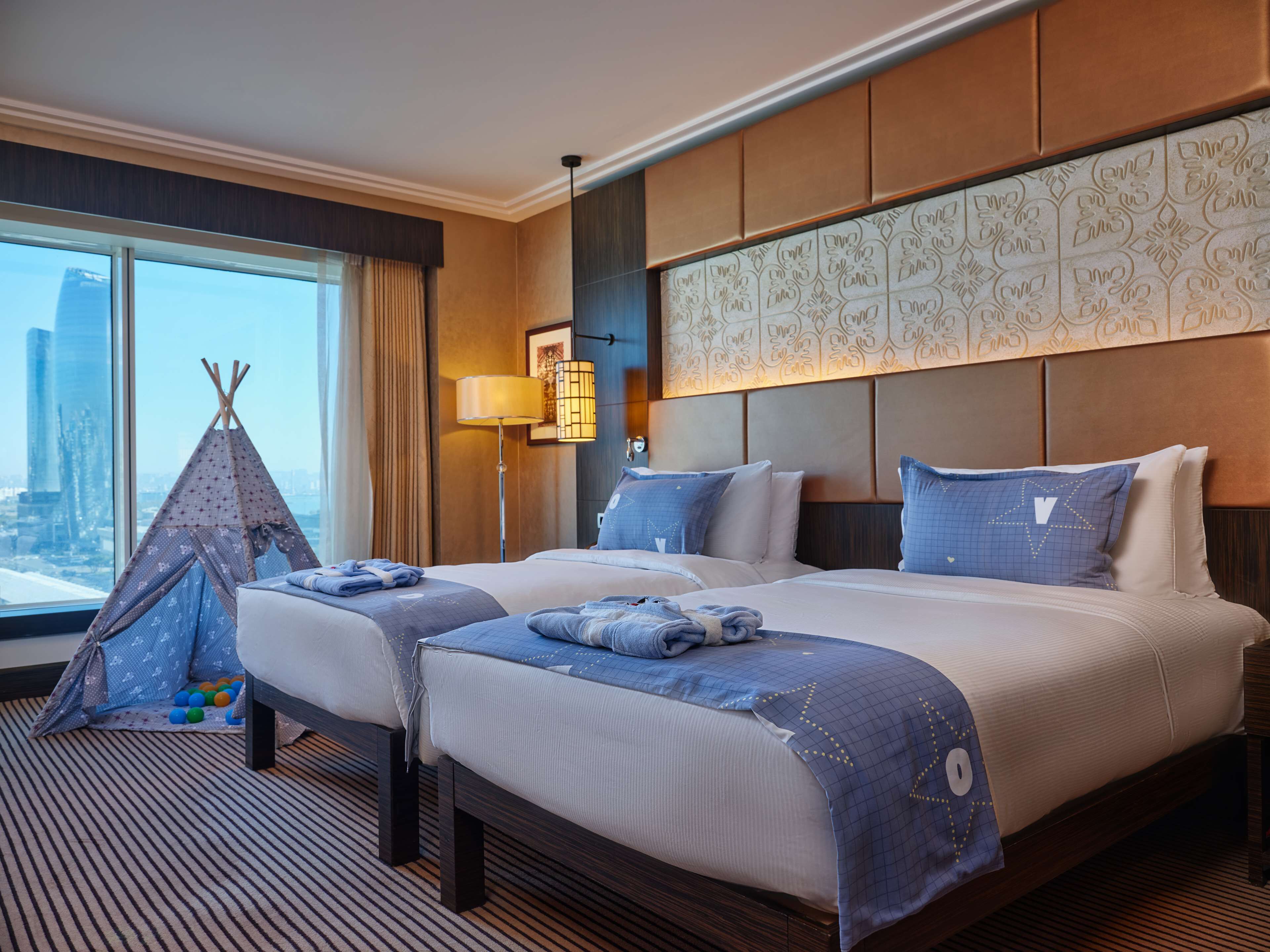 A hotel room with two beds, a city view, a lamp, and a decorative wall panel.