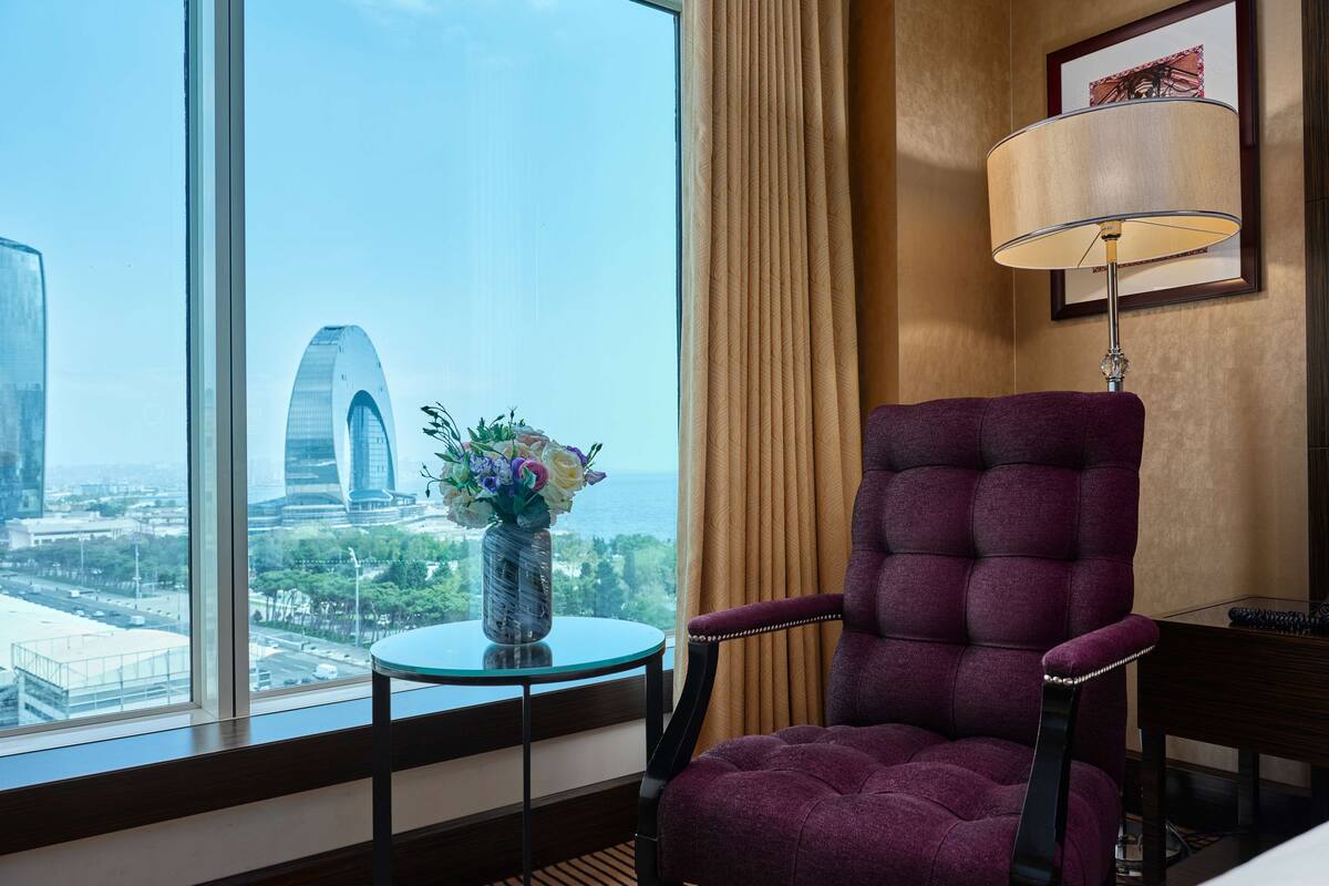 Habitación de hotel con un gran ventanal que ofrece vistas a un paisaje urbano, un sillón morado, una mesita con un florero y una lámpara en una mesita auxiliar.