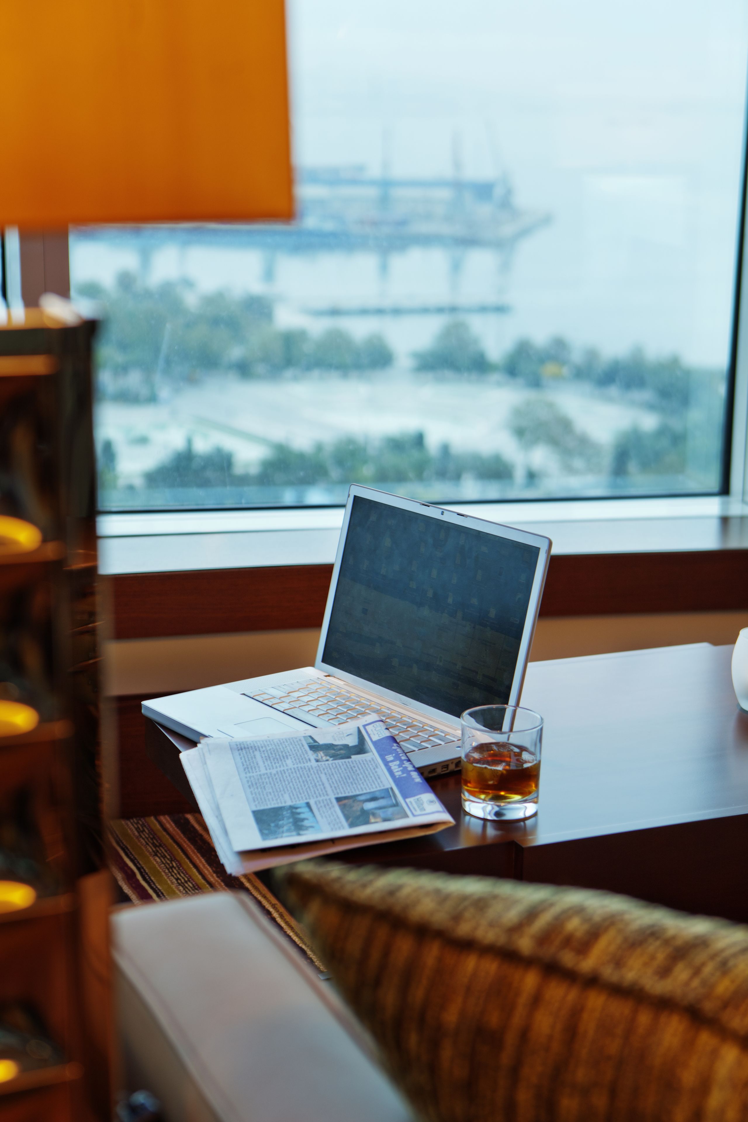 A laptop on a table with a newspaper and a glass of whiskey, near a window with a view of a bridge.