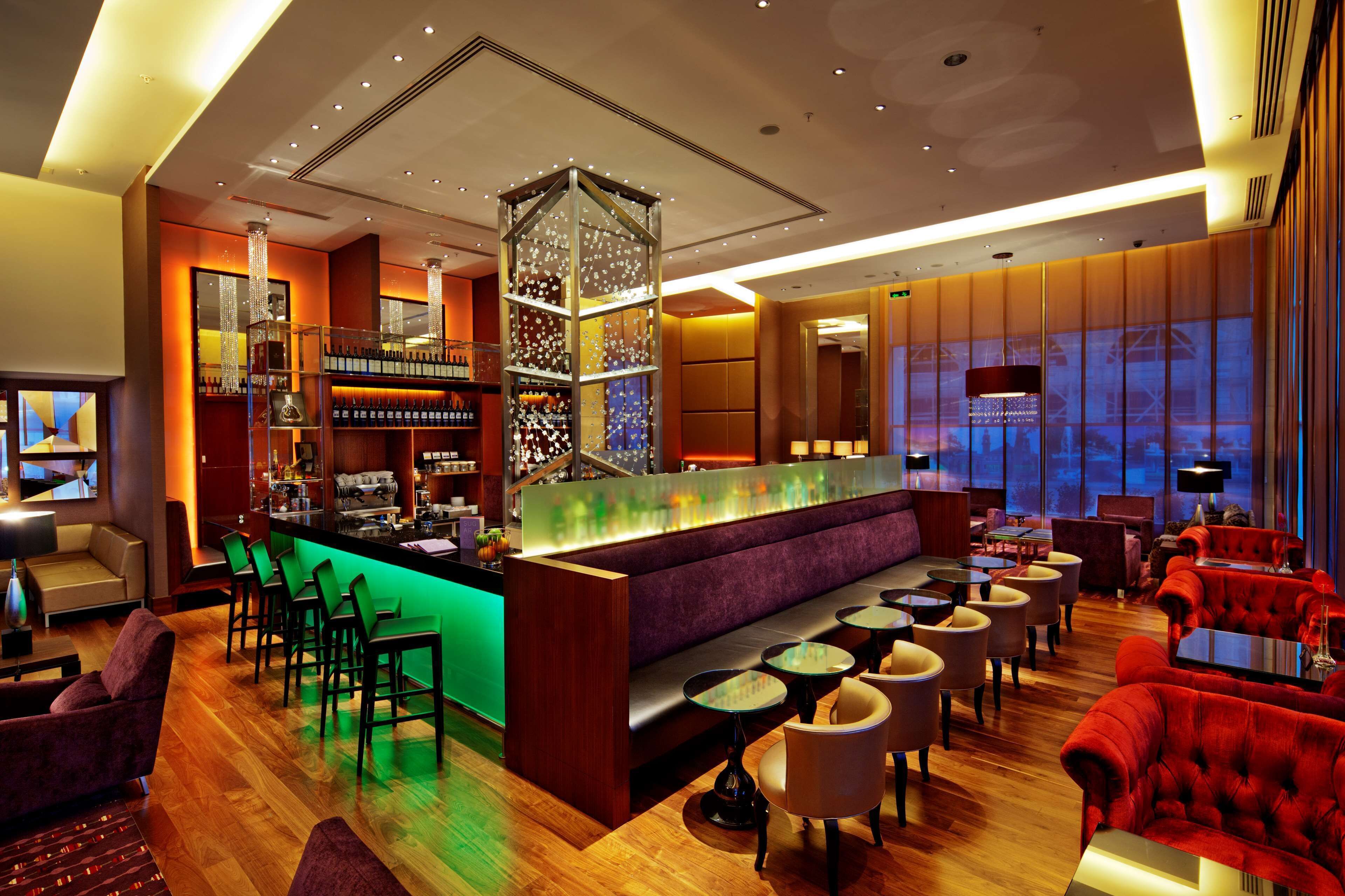 A modern bar area with a central bar, seating arrangements, and a view of the cityscape through large windows.