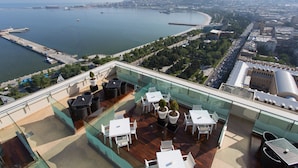 A rooftop terrace with outdoor seating, a view of a coastal cityscape, and a body of water. - Hilton Baku (Baku)
