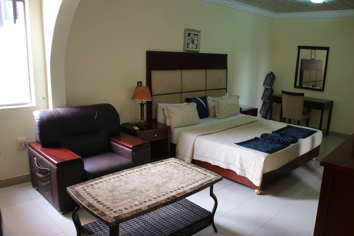 A hotel room with a bed, a desk, a chair, a table, and a lamp.