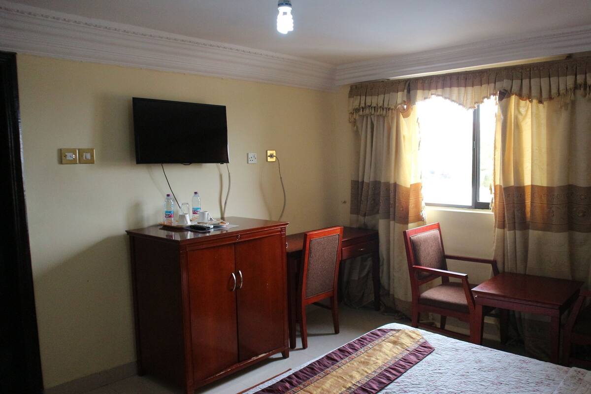 A hotel room with a bed, a desk with a chair, a television mounted on the wall, and a window with curtains.