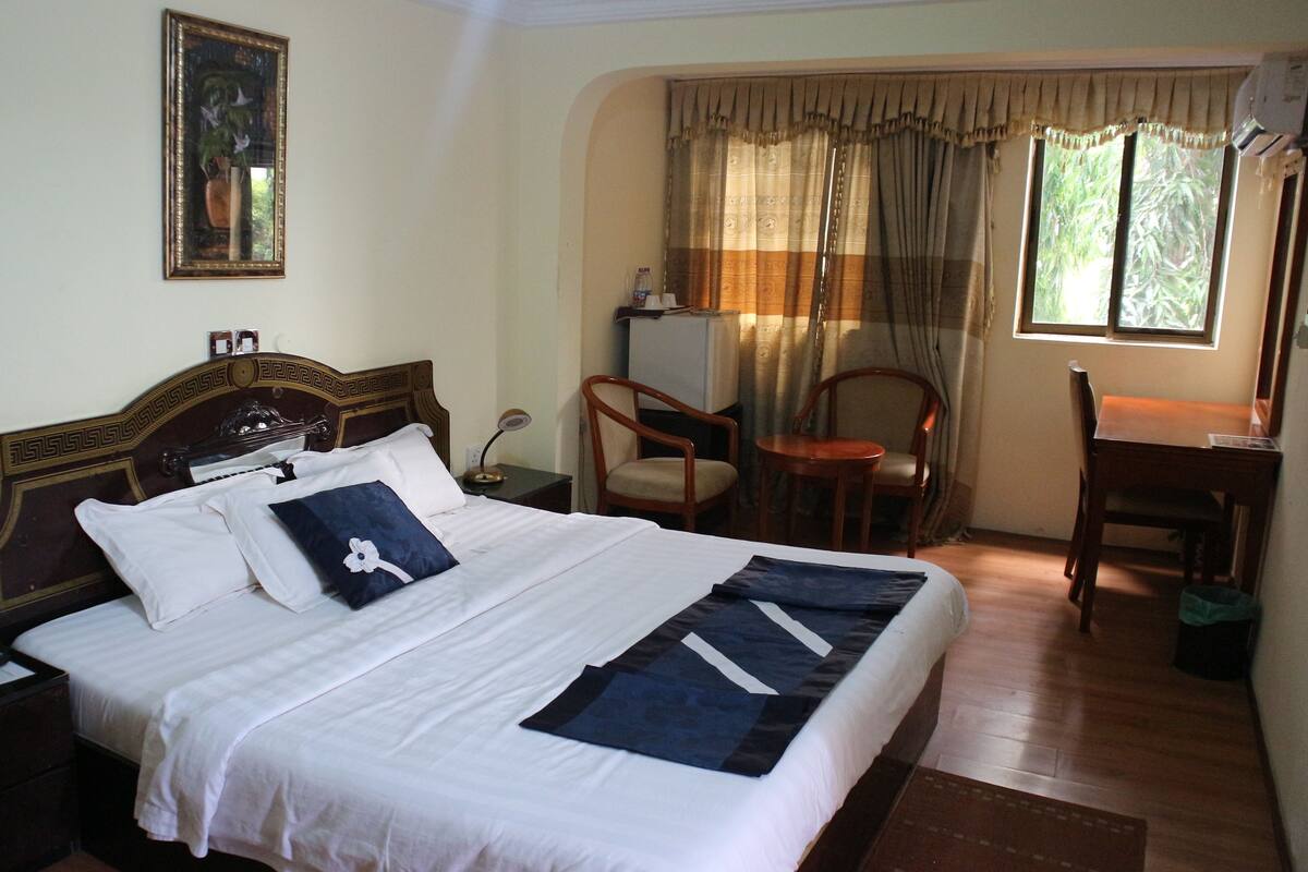 A neatly made bed with white linens and a blue pillow, a wooden nightstand with a phone and papers, a wooden chair, a desk with a lamp, and a window with curtains.