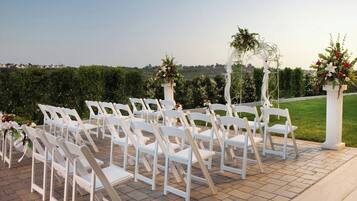 A wedding ceremony setup with white chairs arranged in rows, a floral arch, and a view of a landscaped area.