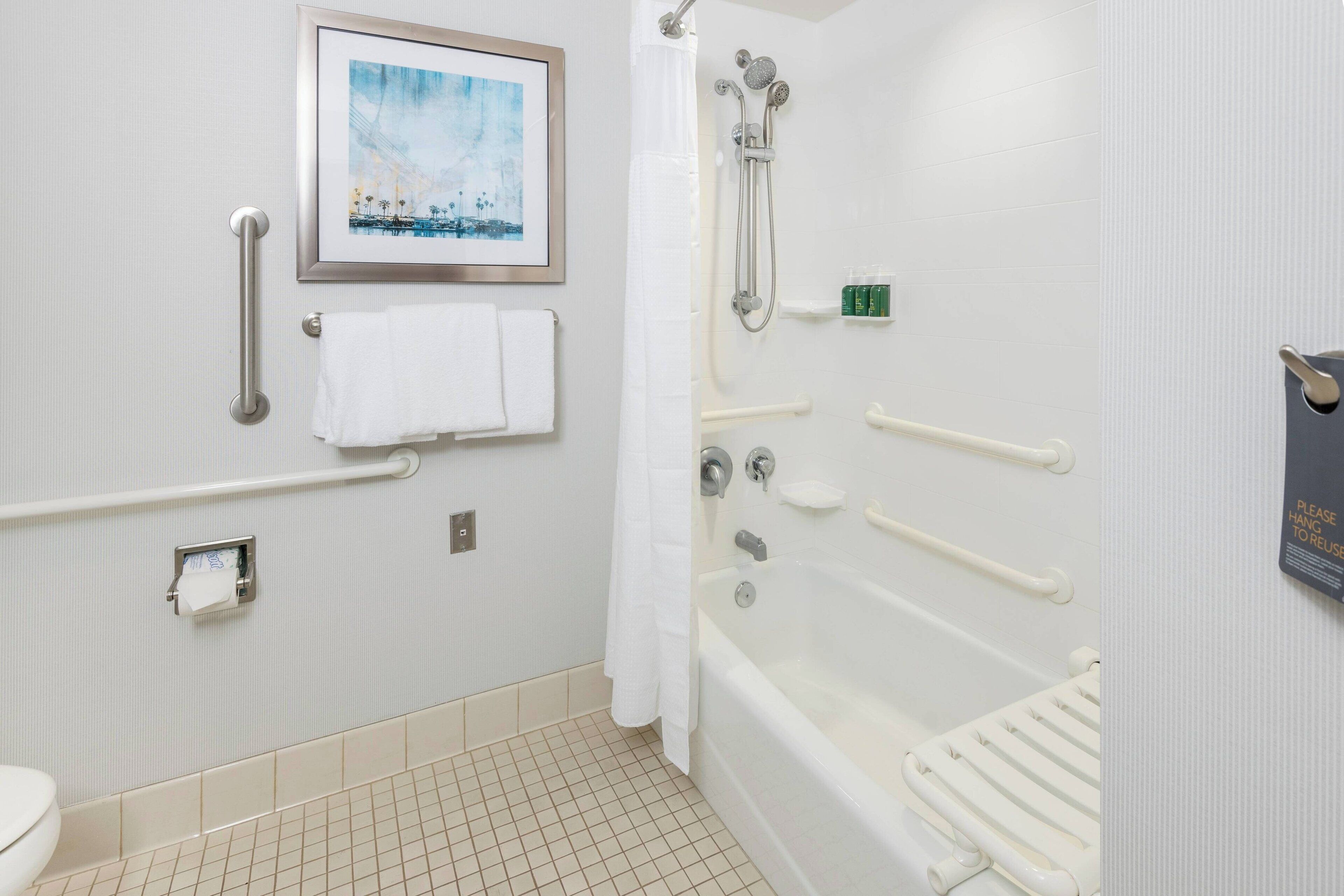 Une salle de bain équipée d’une baignoire, d’une douche, de toilettes, d’une barre d’appui, d’un porte-serviettes et d’un tableau encadré au mur.