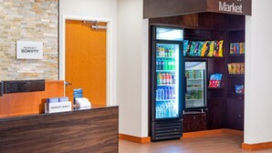 A hotel reception area with a 'Corner Market' sign, a refrigerator stocked with beverages, snacks, and a wooden reception desk. - Fairfield Inn & Suites by Marriott Toronto Mississauga (Mississauga)