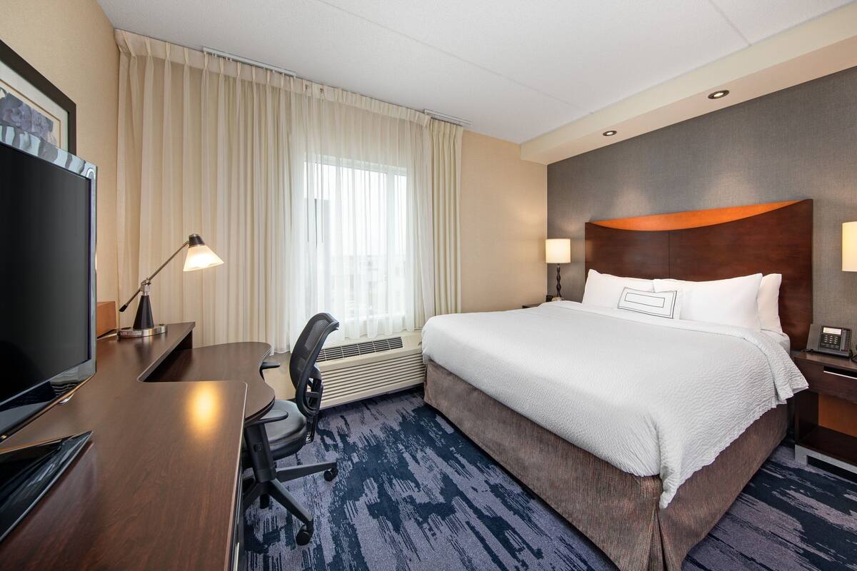 A hotel room with a large bed, a desk with a TV, a chair, a desk lamp, and a telephone.
