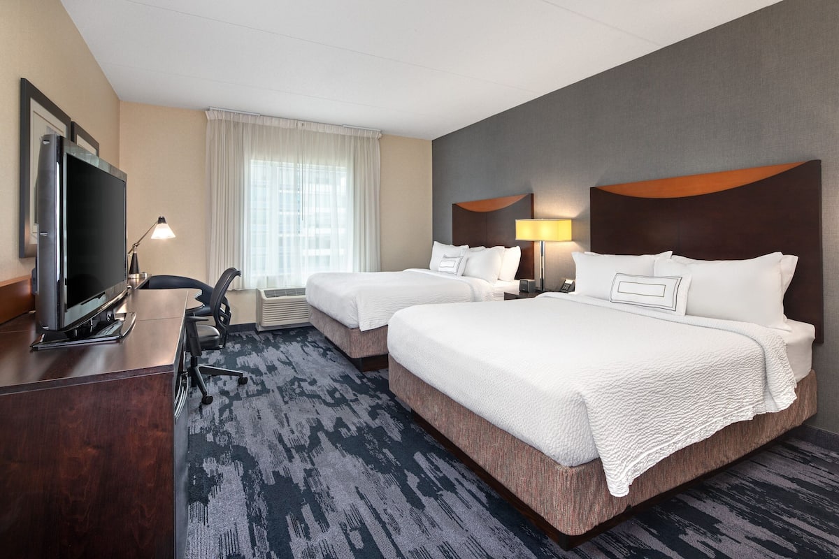 A hotel room with two beds, a desk with a flat-screen TV, a chair, and a large window with curtains.