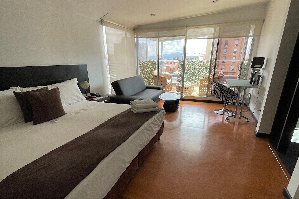 A modern hotel room with a large bed, a sofa, a small table, and a balcony with a city view.