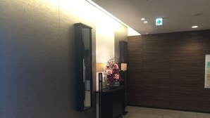 A reception area with a dark wooden wall, a reception desk with a mirror, two lamps, and a floral arrangement. - Meitetsu Inn Kariya (Kariya)