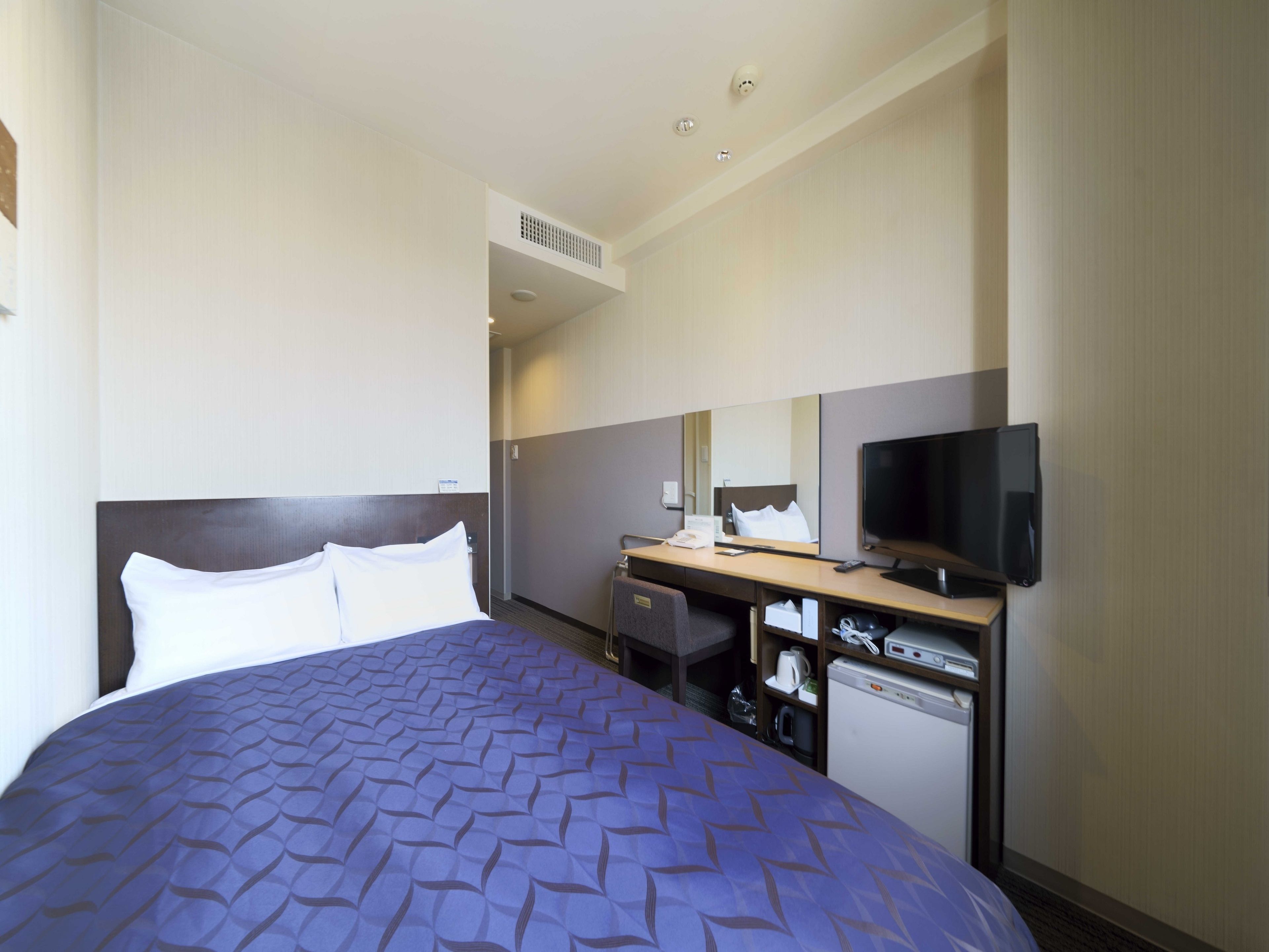A hotel room with a bed, a desk with a television, a mirror, and a small kitchenette.