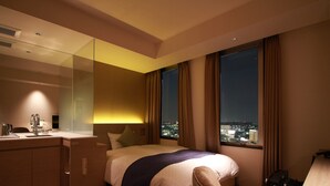 A hotel room with a bed, a desk, a mirror, and a view of the city at night. - Meitetsu Inn Kariya (Kariya)