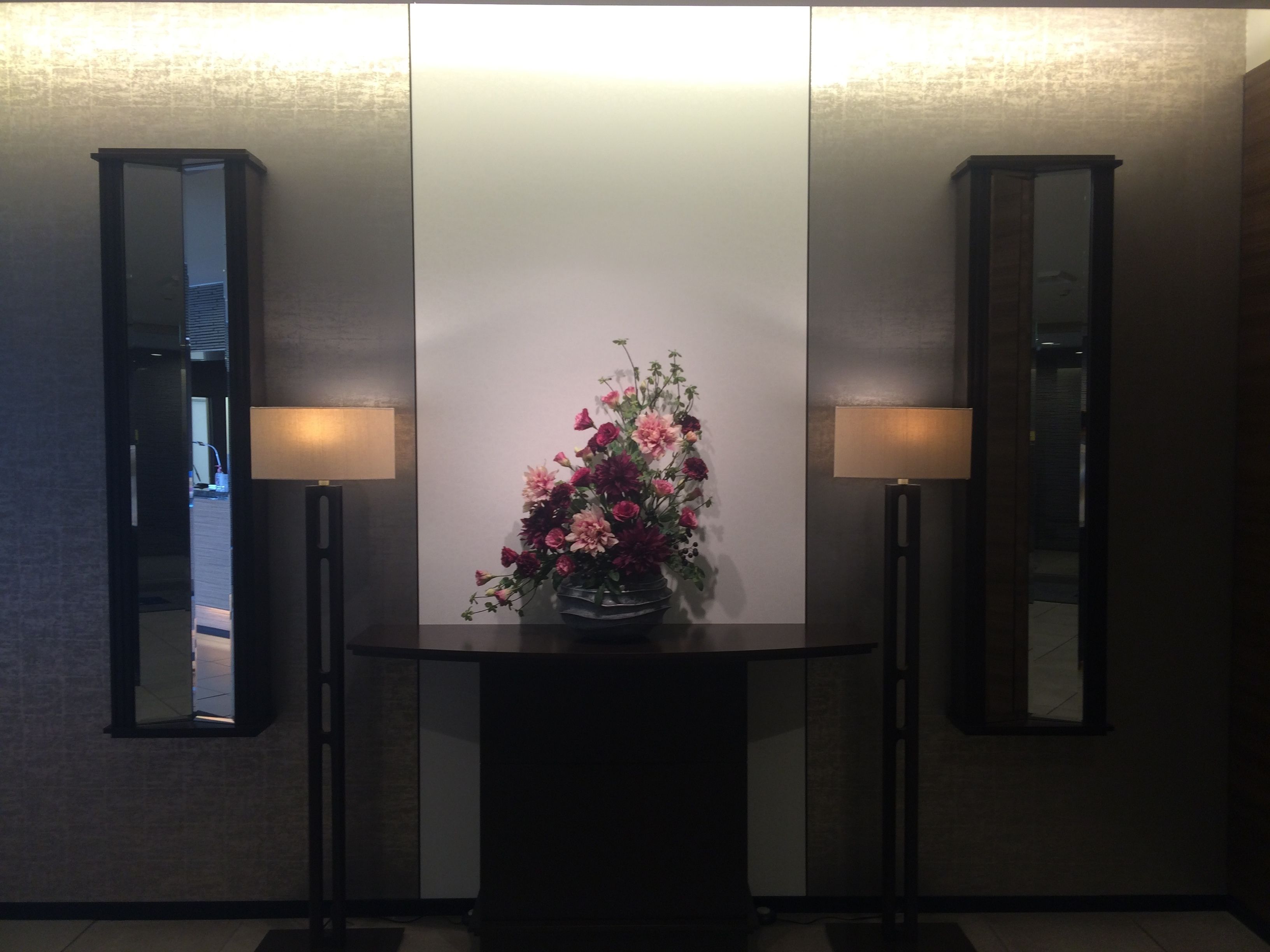 A reception area with a central table displaying a floral arrangement, flanked by two wall-mounted mirrors and two floor lamps.