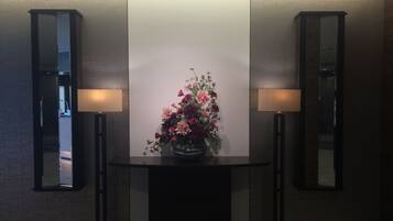A reception area with a central table displaying a floral arrangement, flanked by two wall-mounted mirrors and two floor lamps.