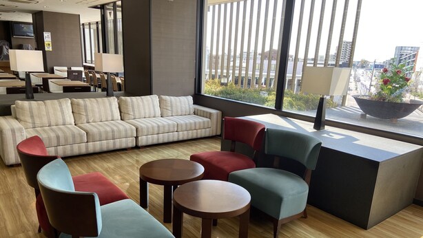 A modern hotel lobby with a sectional sofa, colorful chairs, a small table, and a large window offering a city view. - Meitetsu Inn Kariya (Kariya)