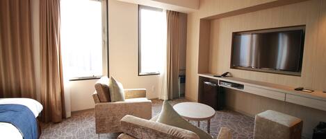 A hotel room with a bed, two armchairs, a small table, a TV, and a desk with a computer.