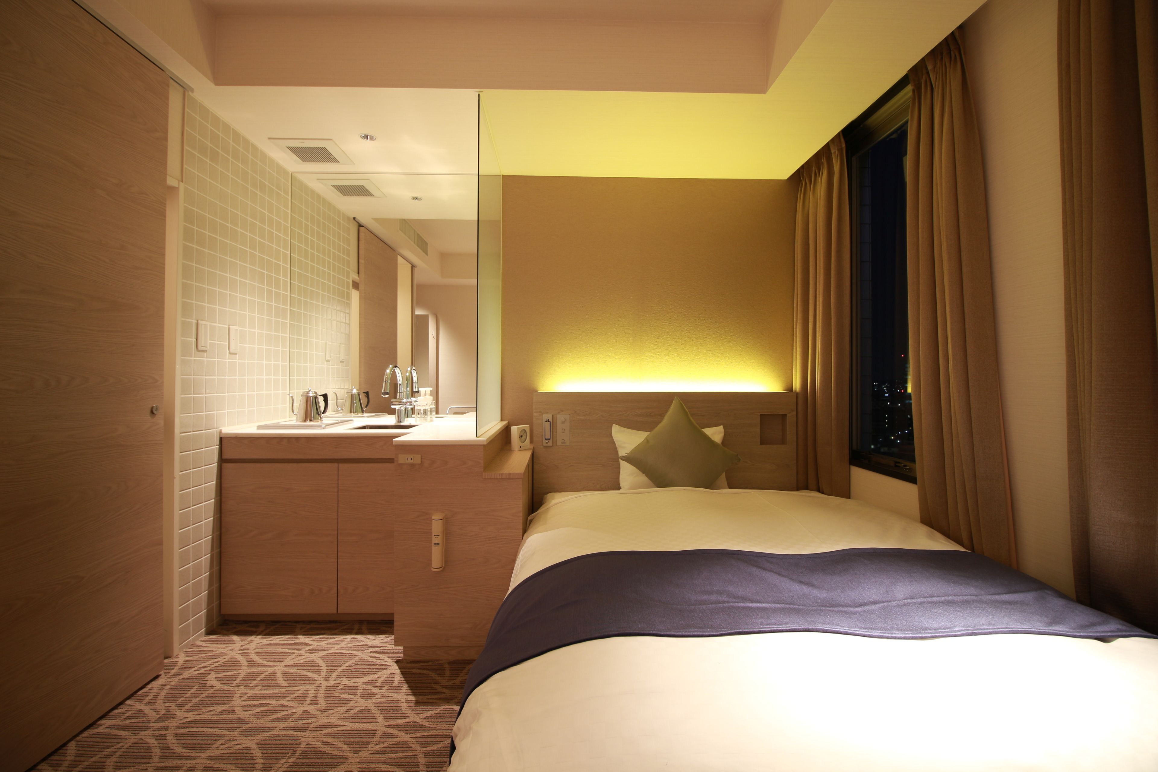 A hotel room with a bed, a nightstand, a mirror, a window with curtains, and a bathroom with a sink and toiletries.