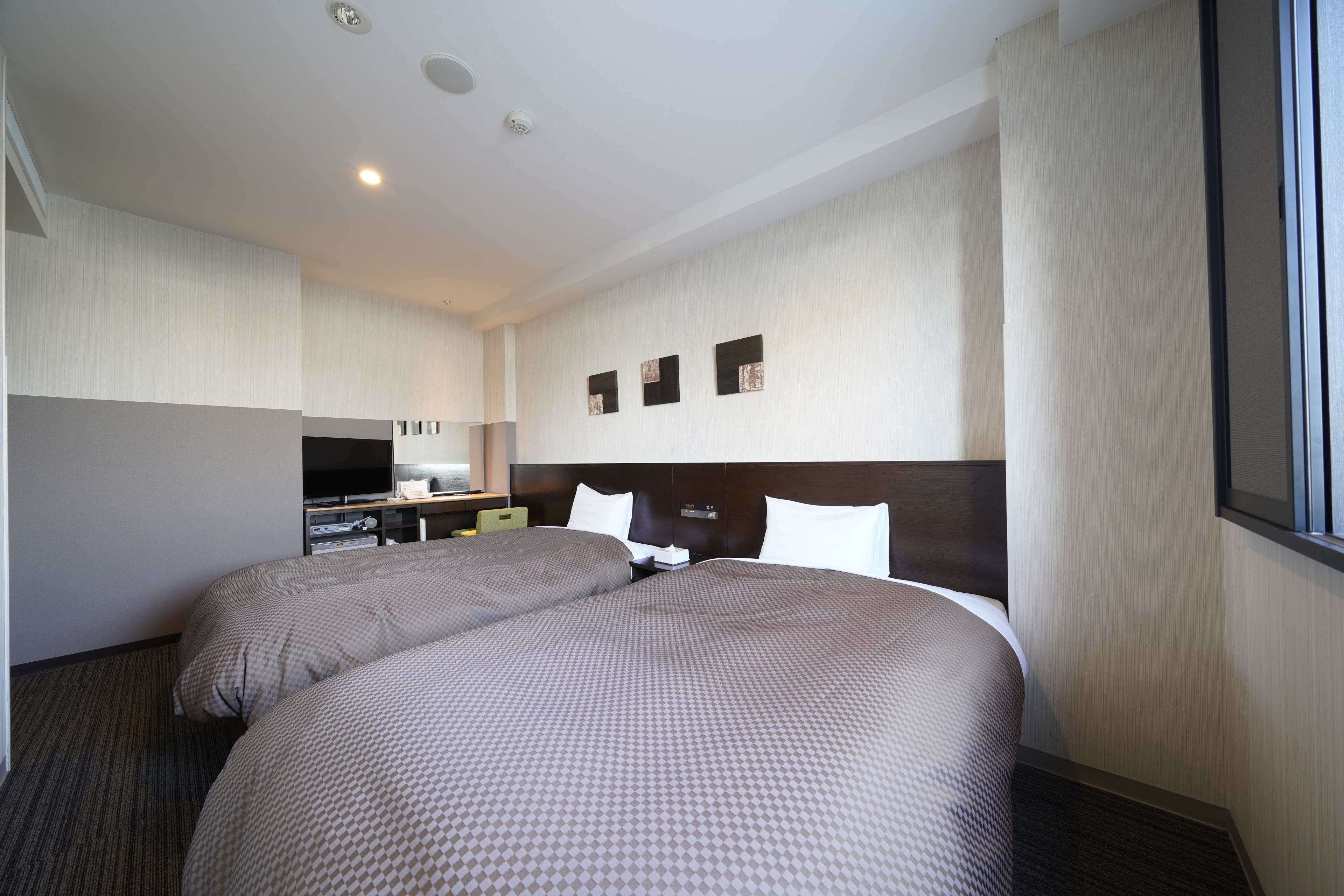 A hotel room with two beds, a desk with a flat-screen TV, a small kitchenette, and a large window.