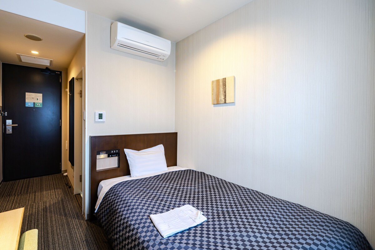 Een hotelkamer met een bed, een wandairconditioner, een deur met een bordje en een klein wandlampje.