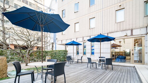 An outdoor patio with round tables and chairs, blue umbrellas, and a building with large windows in the background. - Meitetsu Inn Nagoya Kanayama (Nagoya)