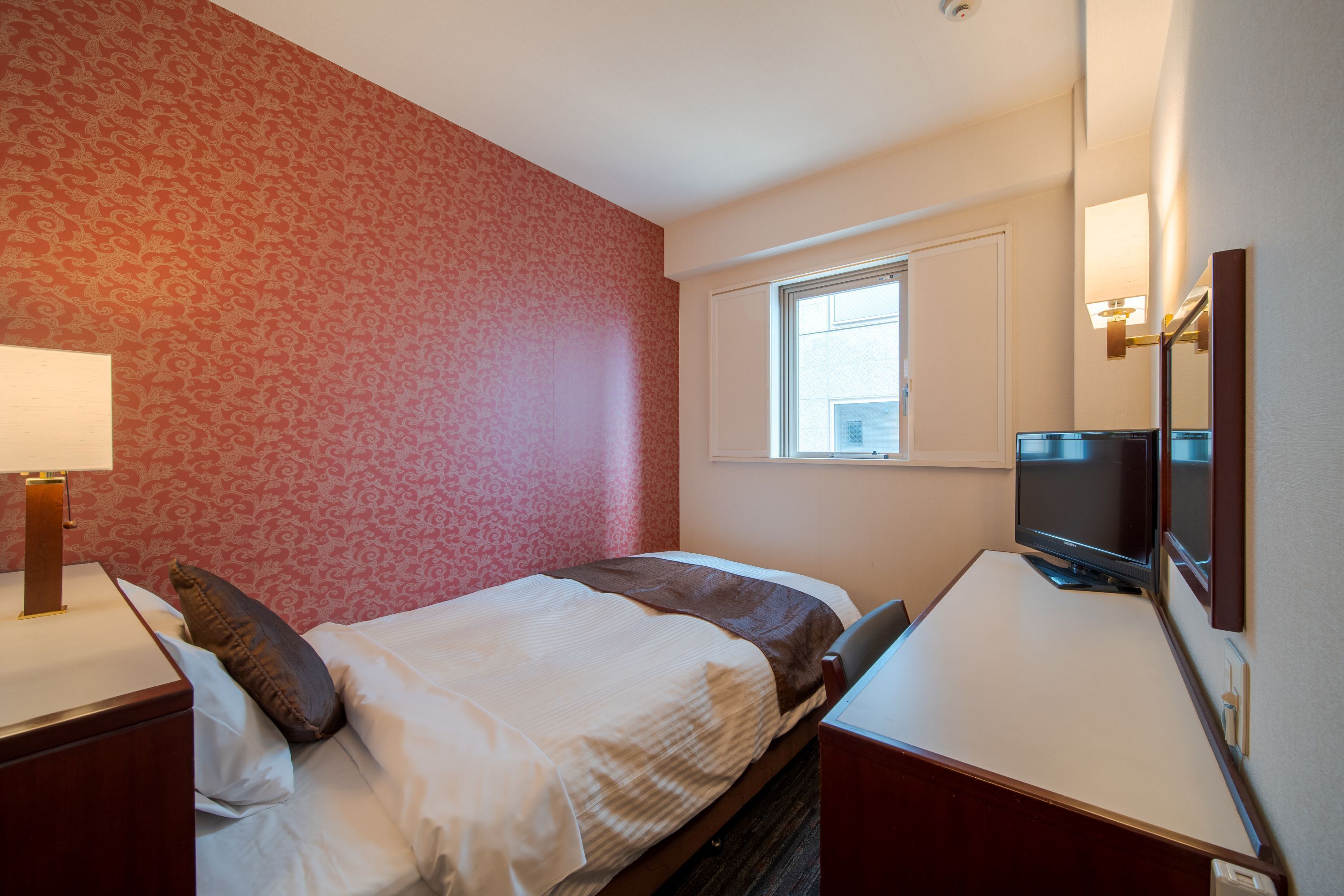 Una habitación de hotel con una cama, un escritorio con televisor, una ventana y una lámpara.