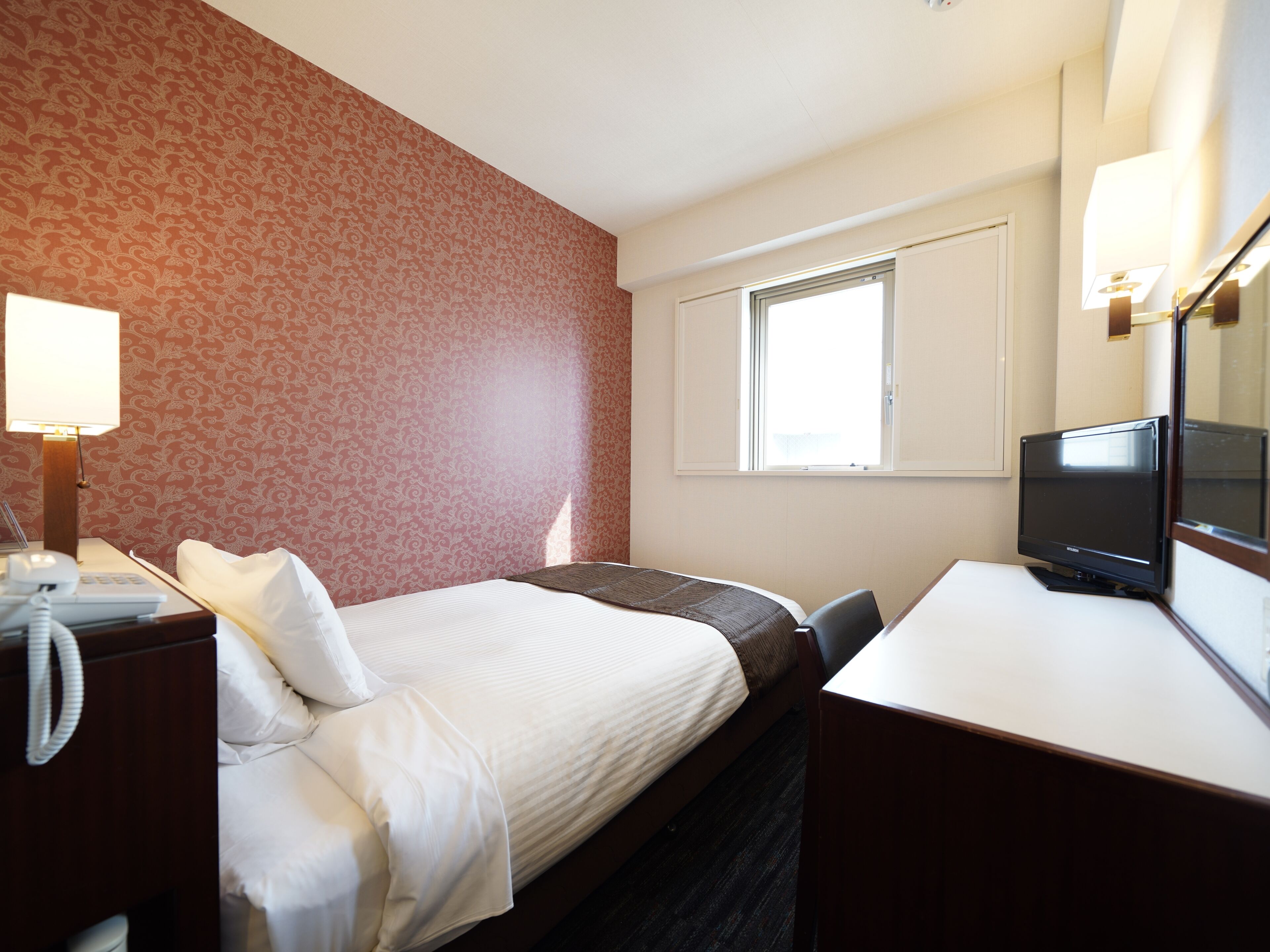 Una habitación de hotel con una cama, una mesita de noche con lámpara, un escritorio con monitor de ordenador y una ventana.