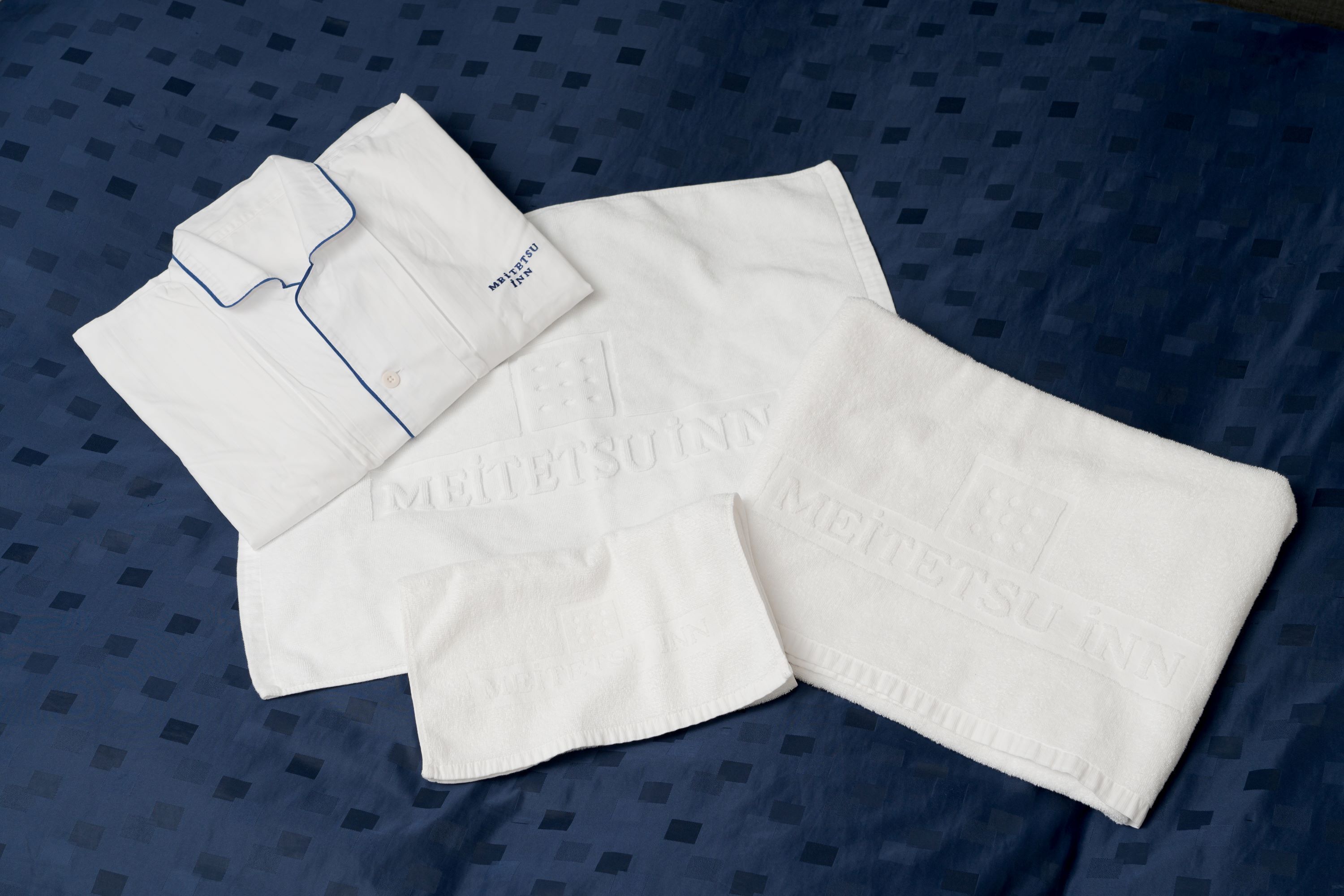 A neatly folded white garment with "MITSU INN" embossed on it, laid on a blue patterned bedspread.