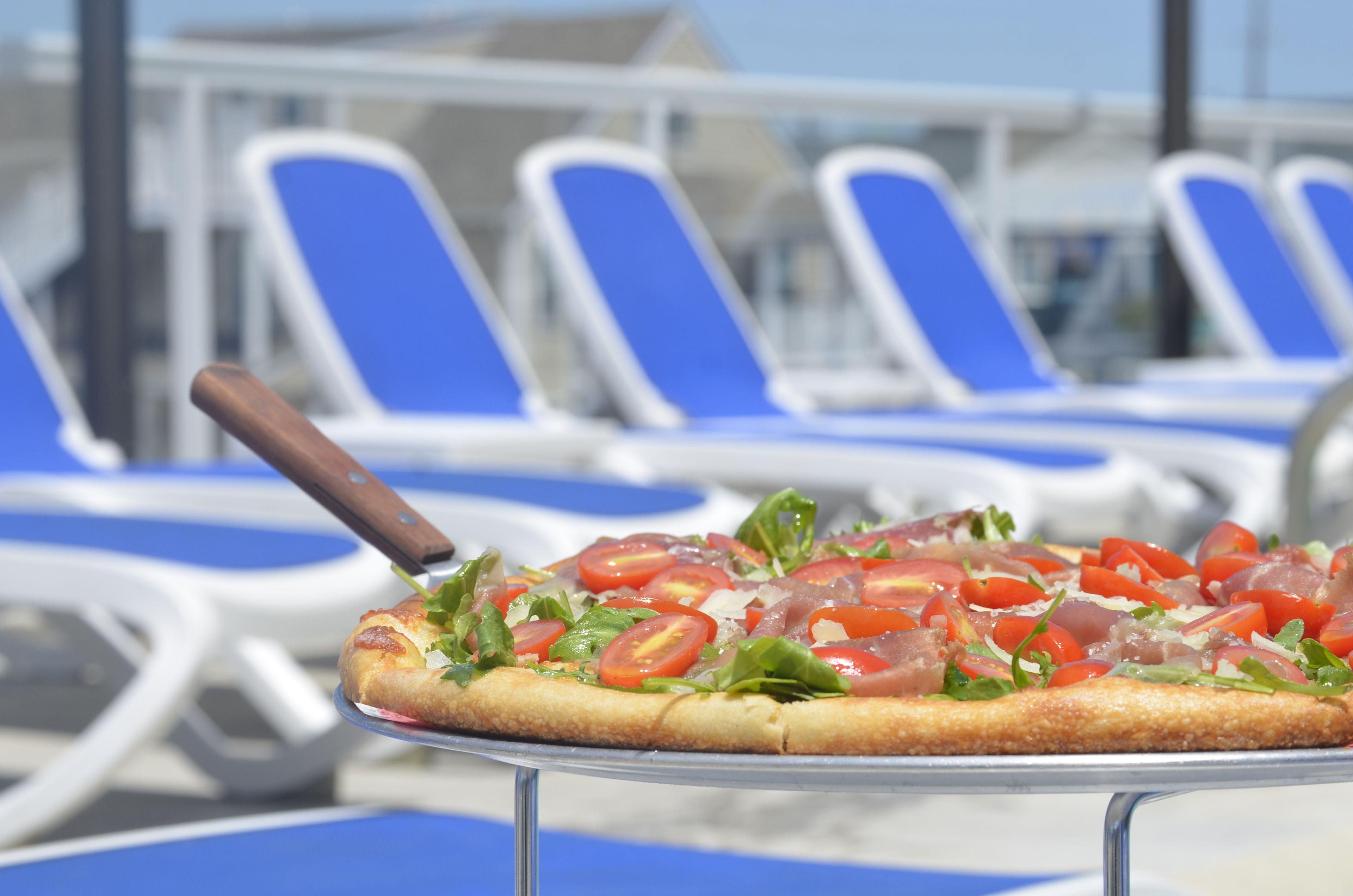 A pizza on a metal stand with a knife on top, surrounded by blue lounge chairs.