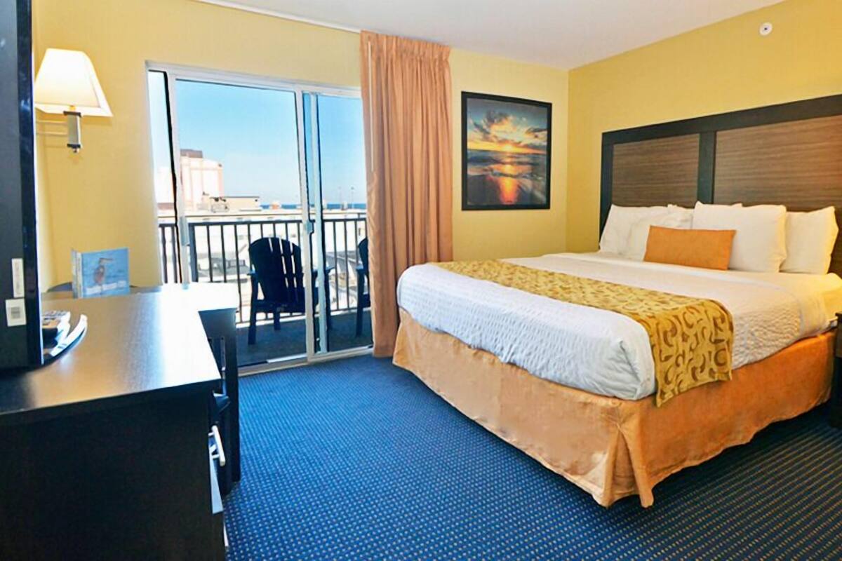 A hotel room with a bed, desk, and chair, a balcony with a view, and a painting on the wall.