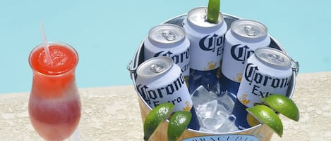 A bucket with Corona beer cans and limes, a glass with a red drink, and a pool in the background.