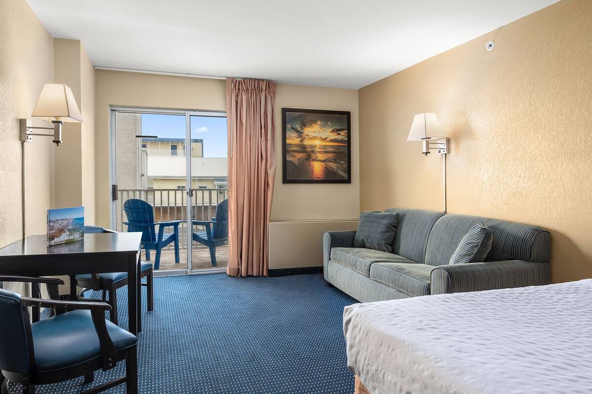 A hotel room with a bed, a sofa, a dining table with chairs, a balcony with a view, and two wall-mounted lamps.