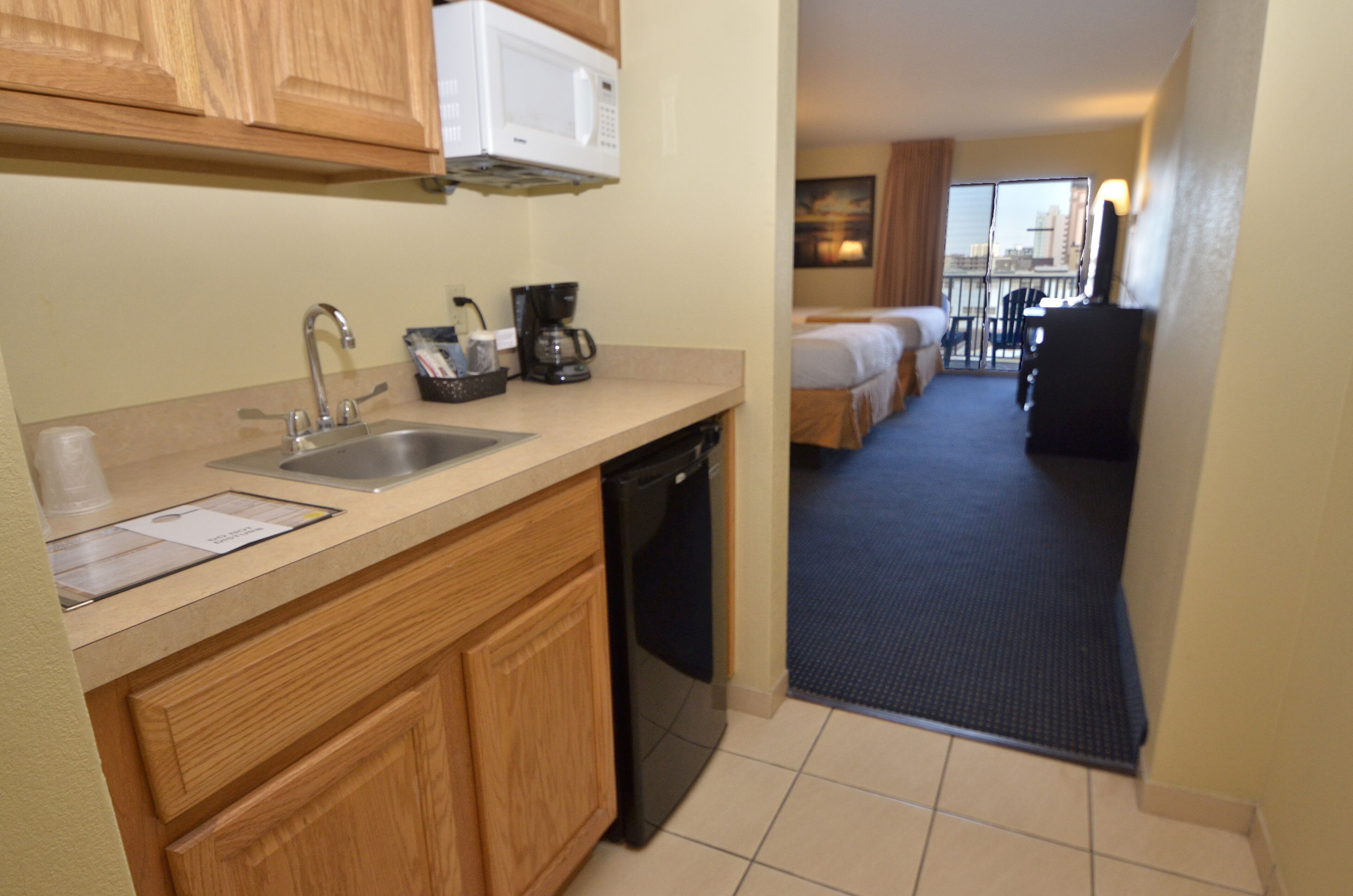 A hotel kitchen with a sink, coffee maker, and microwave. There is a hallway leading to a bedroom with a bed and a chair.