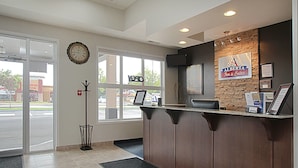 Een receptiegedeelte met een klok, een bord met daarop Alberta Inn & Suites, een kapstok en een raam met uitzicht op een pizzeria.