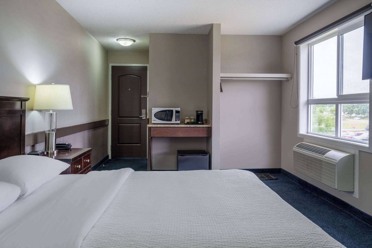 Habitación de hotel con cama, escritorio con microondas, lámpara y ventana con vistas.
